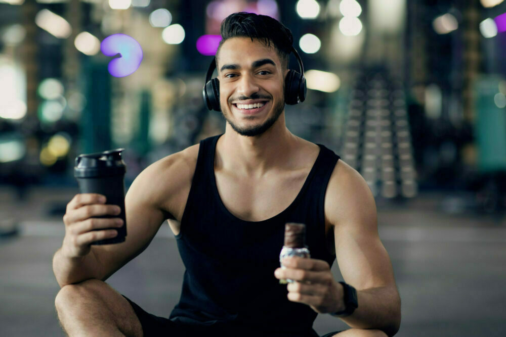 Fitness Nutrition. Happy Muscular Arab Man Holding Sport Shaker And Protein Bar While Relaxing After Training At Gym, Smiling Middle Eastern Male Athlete Having Break In Workout, Enjoying Snacks